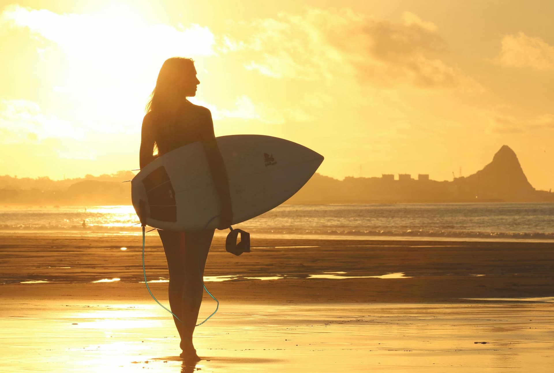 Woman holding a surfboard walking towards the sea with a sunset drop
